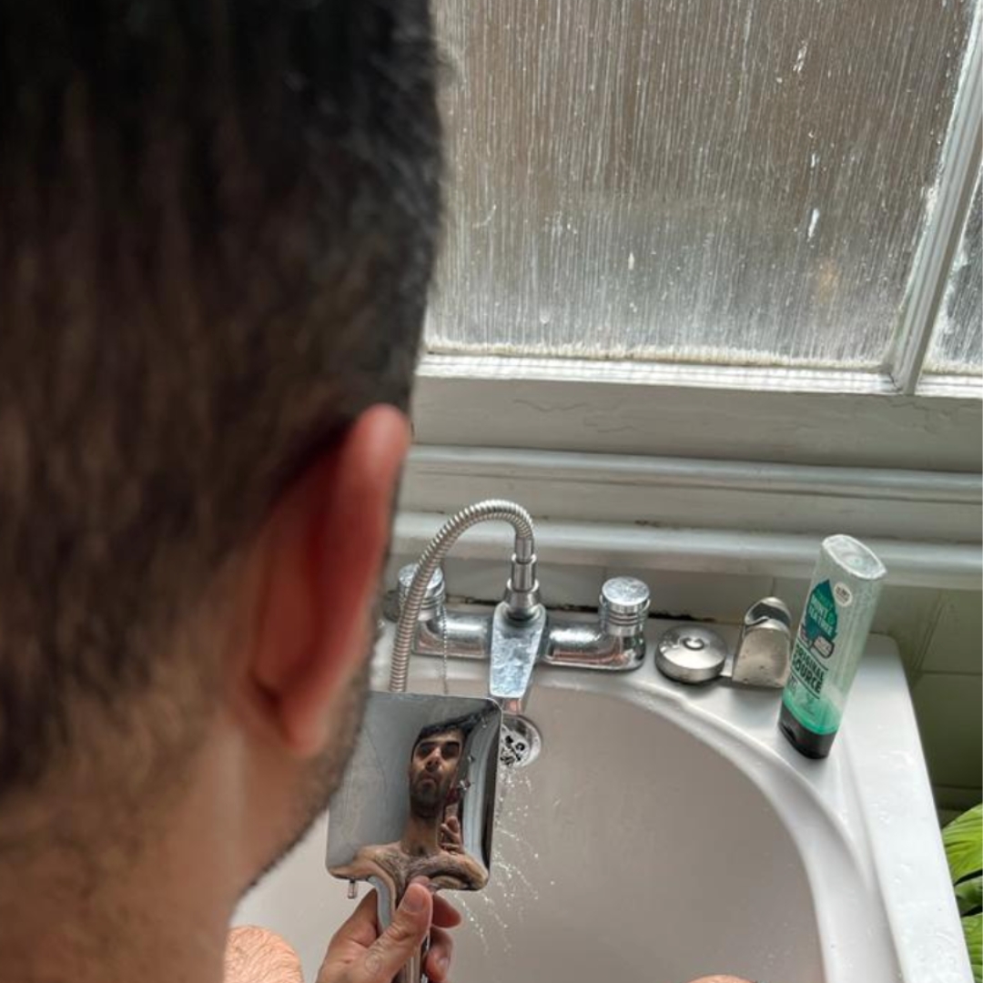 A man stands at the sink, holding a small mirror in which he seems himself reflected, distorted.