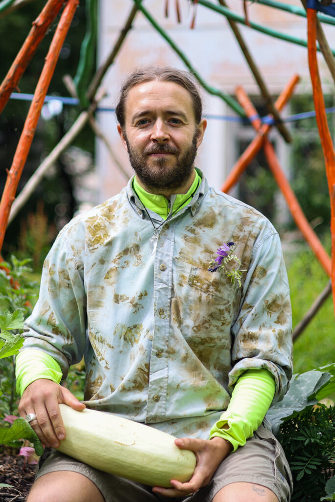 Sean Roy Parker wears a muddy shirt with flowers in the breast pocket. He is in a garden space and holding a large vegetable.